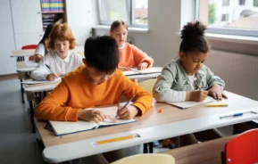 Imagem de cinco crianças do ensino fundamental sentadas em carteiras em uma sala de aula bem iluminada, escrevendo em cadernos. Elas estão concentradas em seus trabalhos, com livros e lápis nas carteiras ao redor.