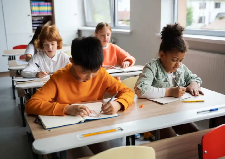 Imagem de cinco crianças do ensino fundamental sentadas em carteiras em uma sala de aula bem iluminada, escrevendo em cadernos. Elas estão concentradas em seus trabalhos, com livros e lápis nas carteiras ao redor.