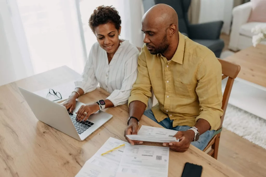Imagem de uma mulher e um homem sentados em uma mesa com um laptop e papéis, discutindo documentos. Enquanto analisam contas e envelopes, eles exploram como calcular a valorização de um imóvel enquanto ela digita no laptop