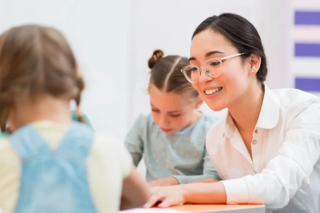 Imagem de uma professora de óculos e camisa branca ajudando duas meninas com os trabalhos escolares em uma sala de aula bem iluminada 