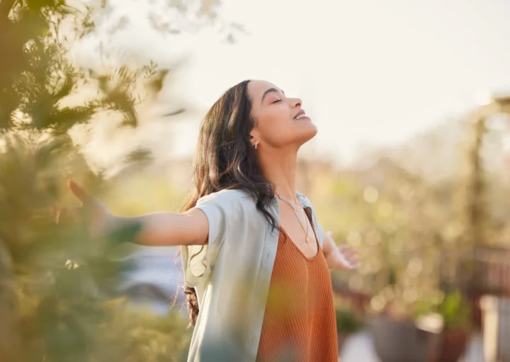 Foto de uma mulher sorridente com os braços abertos, em meio à natureza.