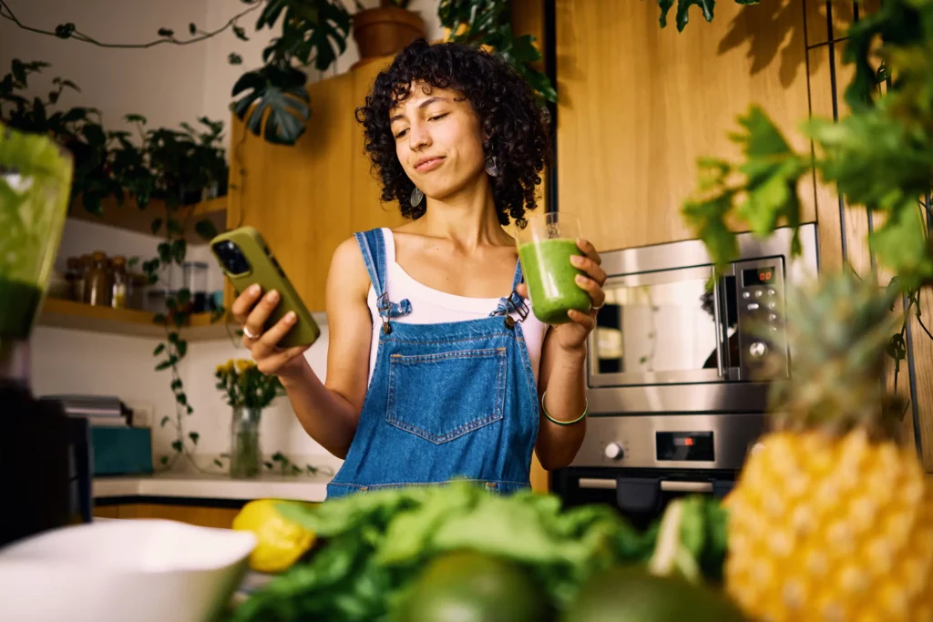 Foto de uma mulher mexendo no celular e tomando suco verde em uma casa repleta de plantas.