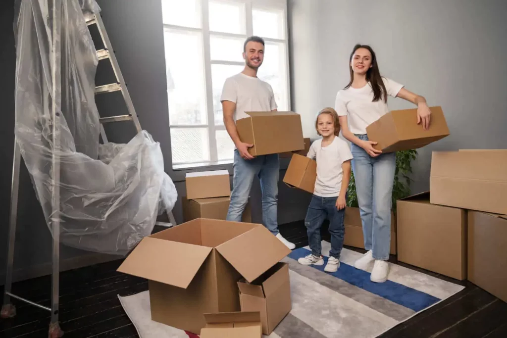 Imagem de uma família jovem de um homem, uma mulher e uma criança com caixas de mudança nas mãos em um cômodo vazio cheio de caixas para ilustrar matéria sobre simulador de consórcio imobiliário