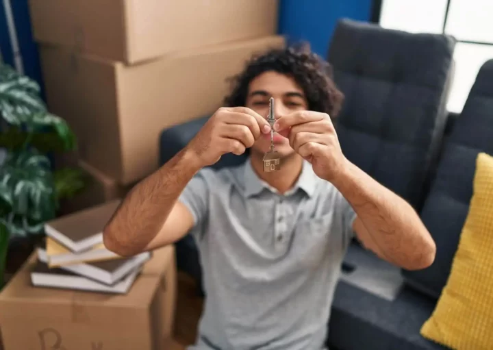 Imagem de um homem sorridente segurando as chaves de um imóvel na altura do rosto dentro de uma sala de estar com um sofá, uma planta e algumas caixas de papelão para ilustrar matéria sobre termo de entrega de chaves