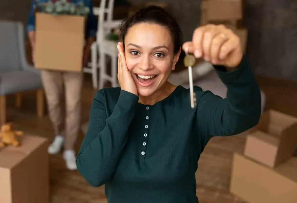 Imagem de uma mulher sorridente segurando as chaves de um imóvel dentro de uma casa enquanto um homem ao fundo carrega uma caixa de papelão para ilustrar matéria sobre o termo de recebimento de chaves