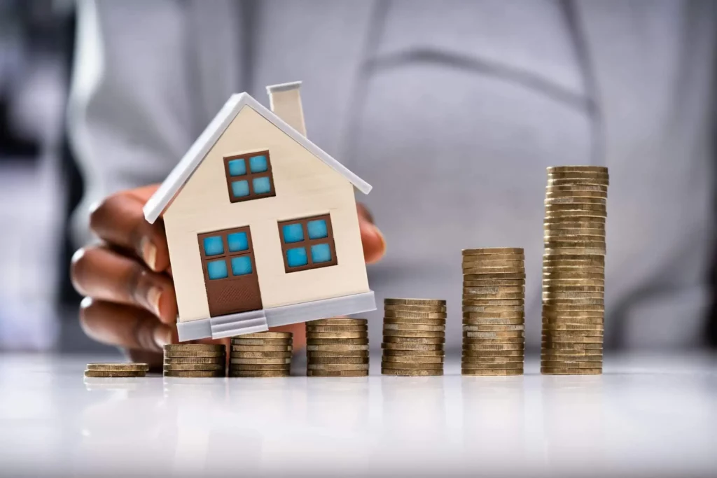 Imagem da mão de uma pessoa segurando uma pequena casa modelo ao lado de pilhas crescentes de moedas, representando visualmente o crescimento do investimento imobiliário ou o aumento do valor da propriedade