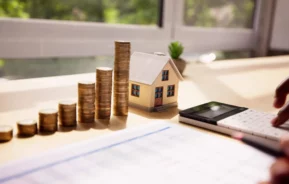 Imagem da mão de uma pessoa usando uma calculadora em uma mesa com moedas empilhadas, uma casa modelo em tamanho miniatura e documentos financeiros. A luz do sol entra pela janela, destacando a vegetação do lado de fora