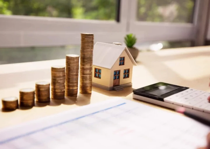 Imagem da mão de uma pessoa usando uma calculadora em uma mesa com moedas empilhadas, uma casa modelo em tamanho miniatura e documentos financeiros. A luz do sol entra pela janela, destacando a vegetação do lado de fora