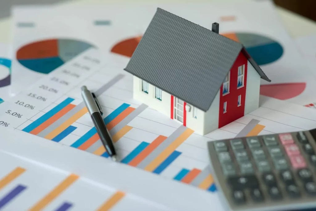 Imagem de uma pequena casa modelo em tamanho miniatura em cima de papéis com o desenho tabelas e gráficos financeiros, com uma caneta e uma calculadora por perto
