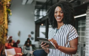 Mulher negra em ambiente empresarial sorrindo e segurando um tablet para ilustrar matéria sobre as cidades com mais empregos no Brasil