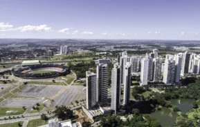Imagem da vista aérea do Parque Flamboyant, em Goiânia, mostra espaços verdes, edifícios altos e modernos, um estádio esportivo e estacionamentos ao redor sob um céu claro