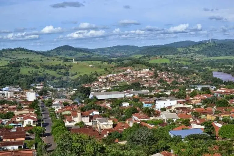 Imagem da vista aérea de Ceres, em Goiás, mostra prédios de telhados vermelhos, colinas verdes e árvores espalhadas sob um céu parcialmente nublado, com um rio ao fundo