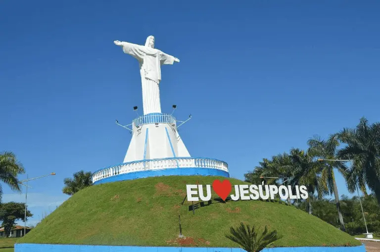 Imagem de uma grande estátua branca do Cristo Redentor fica em uma colina gramada, com uma placa abaixo dizendo ‘eu amo Jesúpolis’, contra um céu azul claro e palmeiras ao fundo