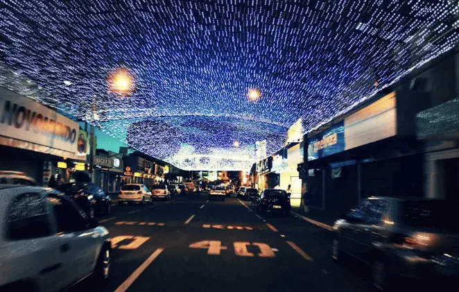 Imagem de uma rua à noite em Catalão, Goiás, com carros estacionados em ambos os lados, iluminados por luzes azuis e brancas que cobrem o céu acima, criando uma atmosfera festiva. Prédios e letreiros de lojas estão alinhados na rua