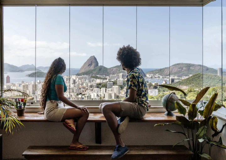 Imagem de duas pessoas sentadas na bancada de um apartamento olhando o Pão de Açúcar, no Rio de Janeiro, pela janela para ilustrar matéria sobre as cidades mais visitadas do Brasil