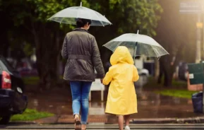 Imagem de uma mulher e uma criança de costas andando em uma faixa de pedestres com guarda chuva na mão para ilustrar matéria sobre as cidades que mais chovem no Brasil