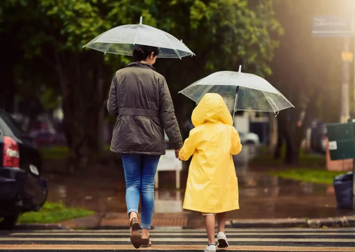 Imagem de uma mulher e uma criança de costas andando em uma faixa de pedestres com guarda chuva na mão para ilustrar matéria sobre as cidades que mais chovem no Brasil