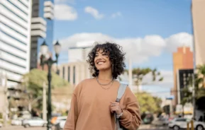 Imagem de uma mulher sorridente com uma mochila nas costas no centro de uma cidade para ilustrar matéria sobre as cidades que mais crescem no Brasil