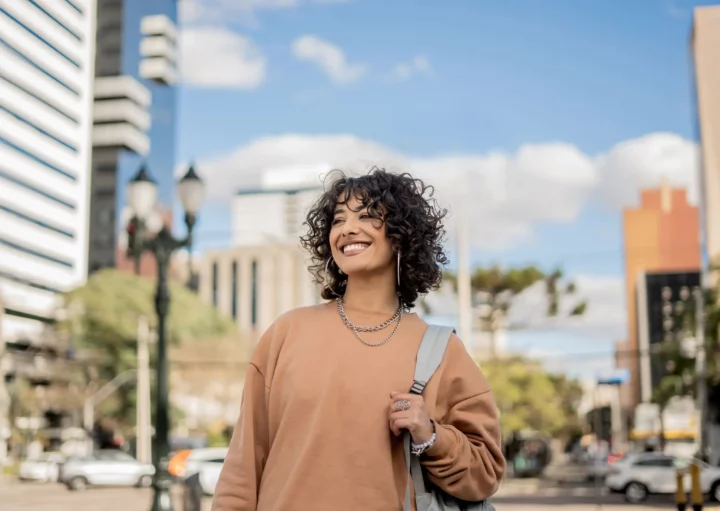 Imagem de uma mulher sorridente com uma mochila nas costas no centro de uma cidade para ilustrar matéria sobre as cidades que mais crescem no Brasil