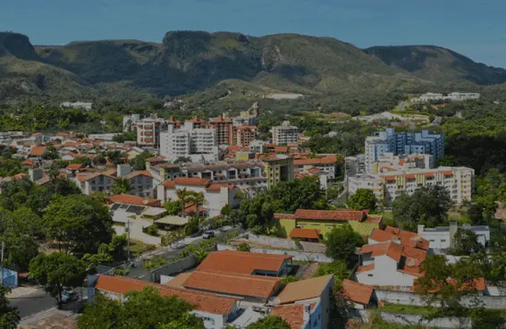 Imagem da vista aérea de Rio Quente, em Goiás, mostra casas de telhado vermelho e prédios de apartamentos, cercada por árvores verdes exuberantes e montanhas sob um céu azul claro