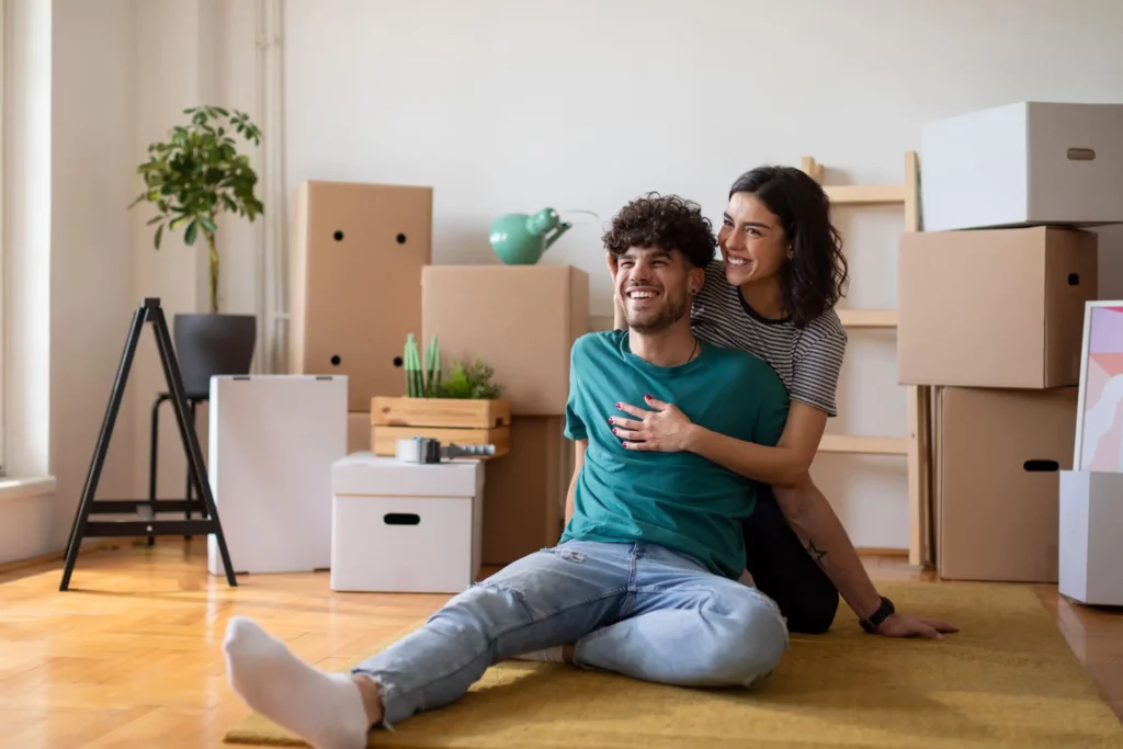 Imagem de um casal composto por um homem e uma mulher sentados no chão de casa com algumas caixas de papelão ao fundo para ilustrar matéria sobre como calcular a fração ideal do imóvel
