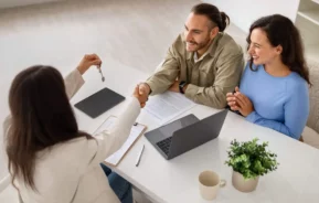 Imagem de um casal formado por um homem e uma mulher sentados na mesa em frente a uma corretora imobiliária que está entregando as chaves de imóvel ao homem enquanto aperta as mãos do comprador em sinal de sucesso para ilustrar matéria sobre contrato de compra e venda de imóvel