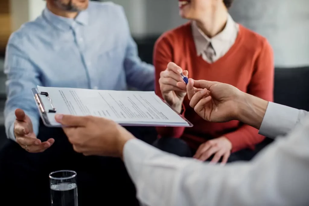 Imagem de uma pessoa de camisa branca entregando uma prancheta com documentos e uma caneta a um casal sorridente, provavelmente pronto para assinar um contrato de compra e venda de imóvel, um deles vestindo uma camisa azul e o outro um suéter laranja.