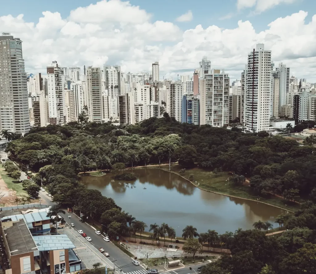 Imagem de um grande parque em Goiânia com um lago e árvores densas é cercado por arranha-céus altos e modernos sob um céu parcialmente nublado. É possível ver carros nas ruas que margeiam o parque