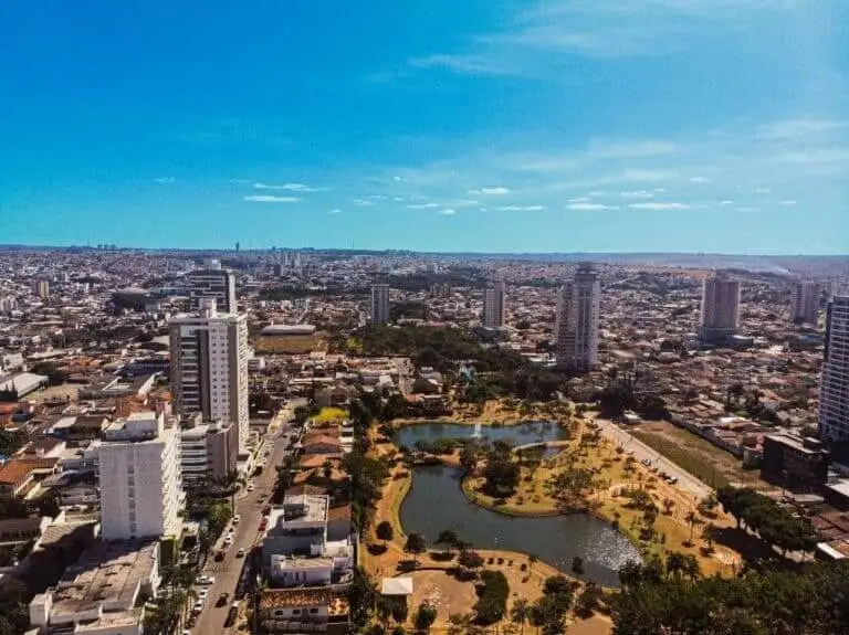 Imagem da vista aérea de Anápolis, em Goiás, mostra prédios altos, um grande parque com lagos, árvores e trilhas para caminhada sob um céu azul claro. A cidade se estende até o horizonte ao fundo