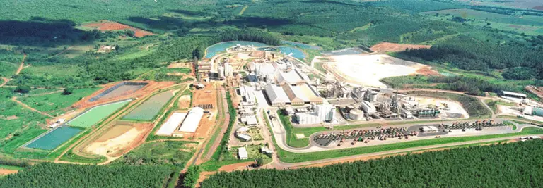 Imagem da vista aérea de um grande complexo industrial cercado por campos verdes, florestas e lagoas de água em Ouvidor, Goiás, com vários edifícios, áreas de armazenamento e estradas visíveis dentro da instalação