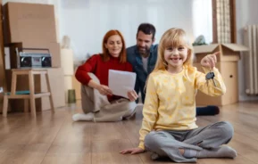 Imagem de uma criança sentada no chão segurando a chave de uma casa enquanto os pais estão atrás sentados ao redor de caixas de papelão analisando um contrato para ilustrar matéria sobre o programa Minha Casa, Minha Vida
