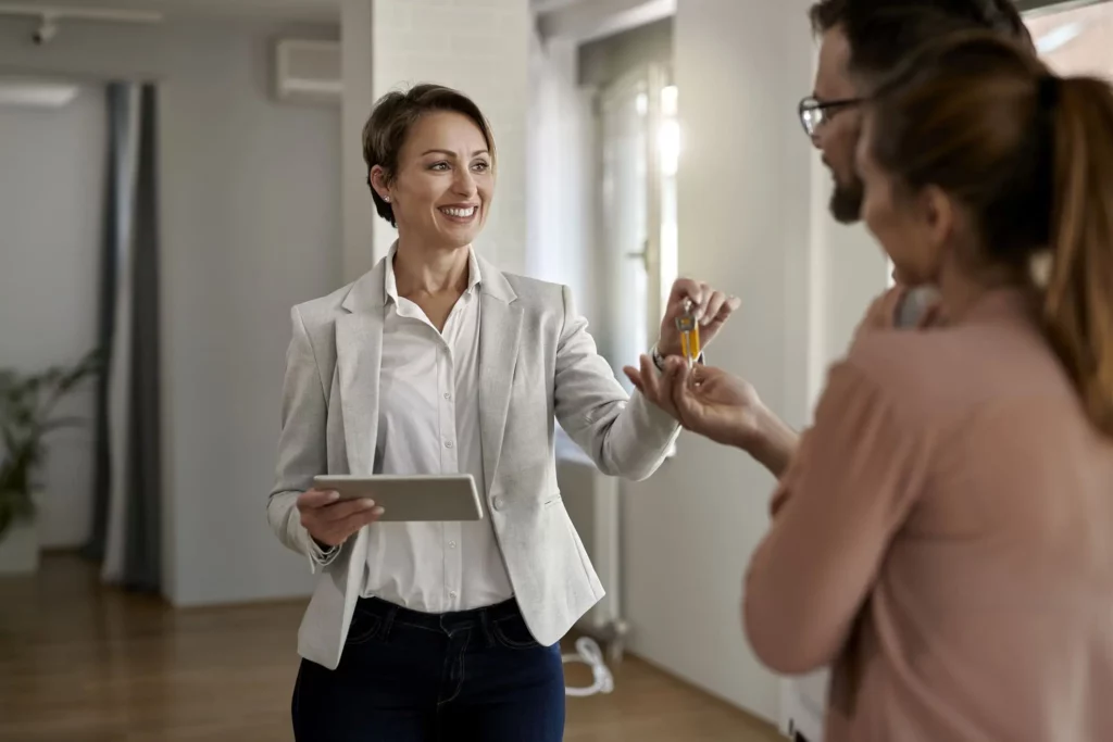 magem de uma mulher sorridente entregando uma chave a um casal composto por um homem e uma mulher para ilustrar matéria sobre quanto devo ganhar para morar de aluguel