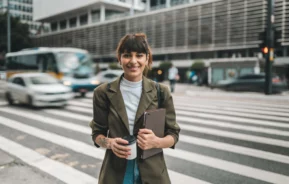 Imagem de uma adulta em frente a uma faixa de pedestres sorrindo e vestindo roupas formais para ilustrar matéria sobre o ranking das cidades mais ricas do Brasil