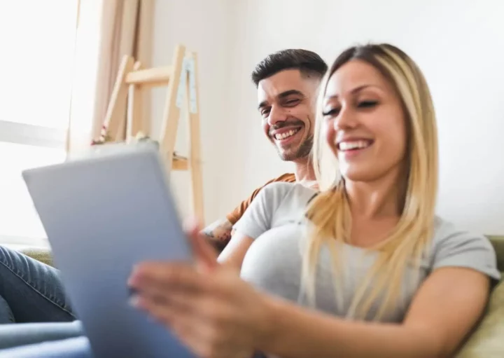 Imagem de um casal sorridente sentado junto em um sofá, olhando para a tela de um tablet para ilustrat matéria sobre o que é tabela SAC. A mulher segura o tablet enquanto o homem se senta ao lado dela. Ambos parecem relaxados e felizes em uma sala iluminada e aconchegante.