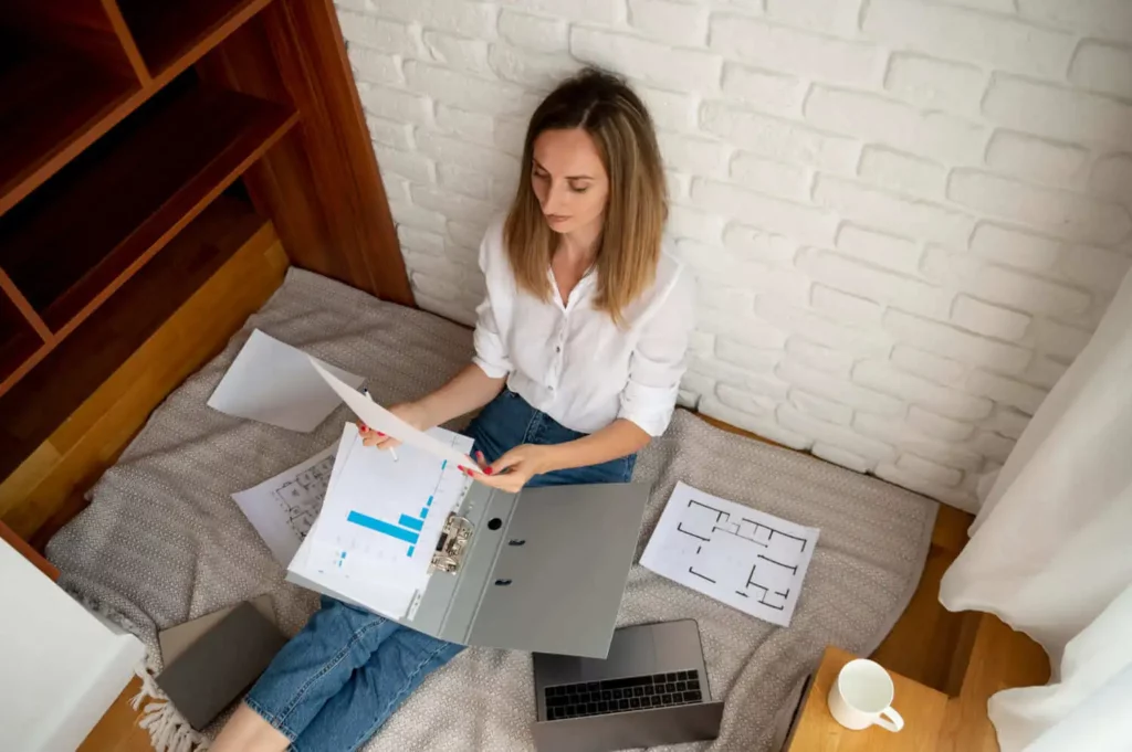 Imagem de uma mulher sentada no chão com papéis, um fichário e um laptop, enquanto analisa documentos. Ela está vestindo uma camisa branca e calça jeans, cercada de papéis em uma sala aconchegante e bem iluminada.