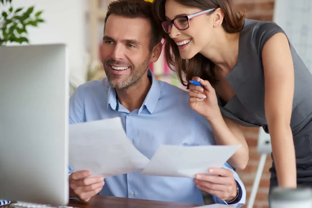 Imagem de um homem e uma mulher sorrindo enquanto olham para um monitor de computador enquanto a mulher, de óculos, se inclina sobre seu ombro com uma caneta na mão.