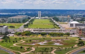 Imagem da vista aérea do Eixo Monumental de Brasília, com modernos prédios governamentais, amplos gramados verdese a paisagem urbana ao longe sob um céu parcialmente nublado para ilustrar matéria sobre o valor do m² em Brasília