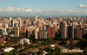 Foto que ilustra matéria sobre a cidade mais populosa do Paraná mostra a capital Curitiba vista do alto, com uma parte arborizada em primeiro plano, muitos prédios no centro da imagem e um céu azul com pucas nuvens ao fundo. (Foto: Renato Soares | Mtur)