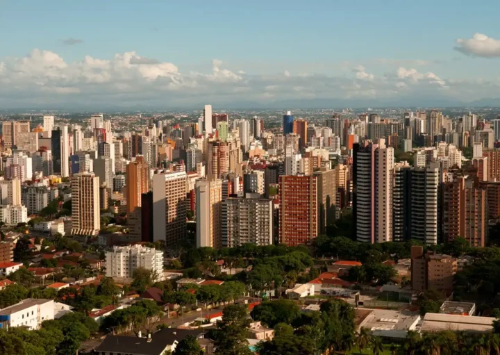 Foto que ilustra matéria sobre a cidade mais populosa do Paraná mostra a capital Curitiba vista do alto, com uma parte arborizada em primeiro plano, muitos prédios no centro da imagem e um céu azul com pucas nuvens ao fundo. (Foto: Renato Soares | Mtur)