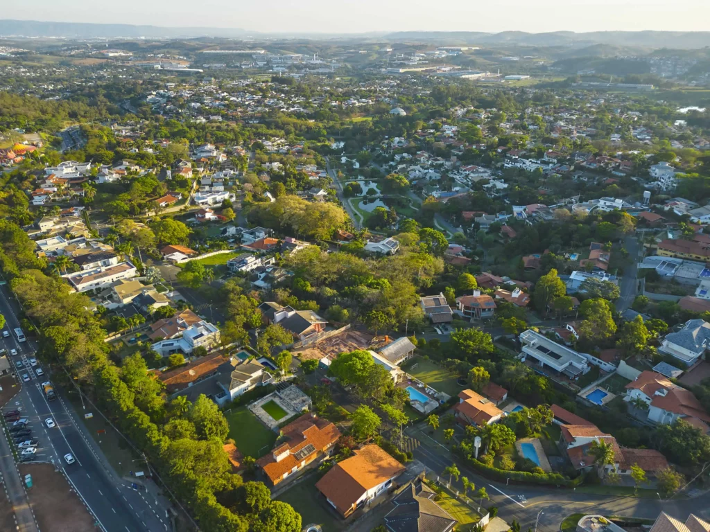 Imagem da vista aérea de um bairro de São Paulo, no Brasil, mostra ruas arborizadas, casas com telhados de telhas vermelhas, piscinas e espaços verdes sob um céu claro
