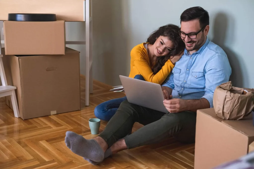 Imagem de um homem e uma mulher sentados no chão de um imóvel olhando para a tela de um notebook com algumas caixas de papelão ao lado para ilustrar matéria sobre como funciona o Casa Paulista