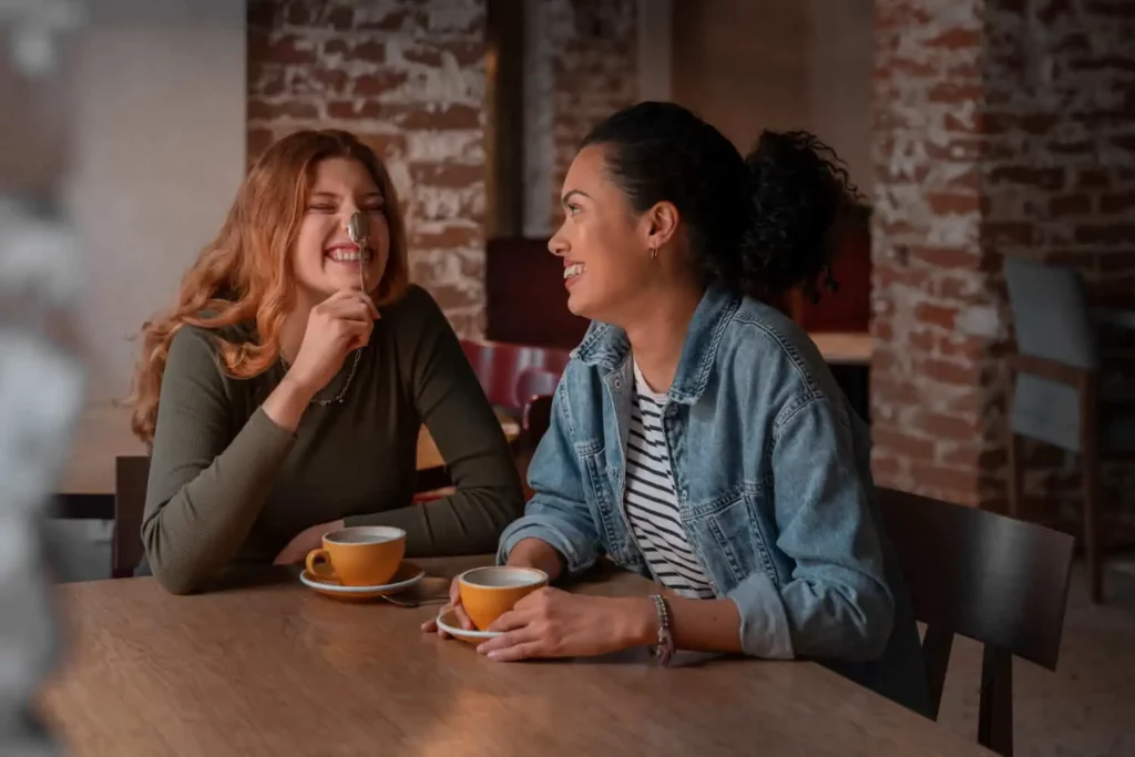 Imagem de duas mulheres sentadas em uma mesa de madeira em uma cafeteria, sorrindo e rindo juntas enquanto seguram xícaras de café.