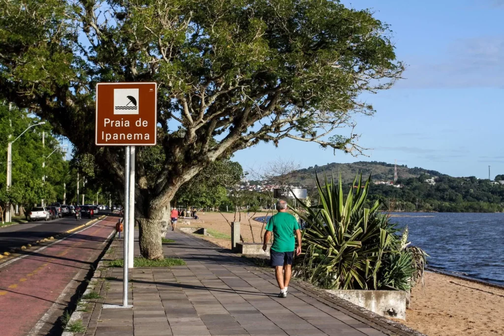 Imagem de um homem de camisa verde caminhando por um caminho pavimentado perto de árvores e plantas, com uma placa marrom da Praia de Ipanema, na Zona Sul de Porto Alegre. Os carros se alinham na rua e as colinas se erguem ao fundo.