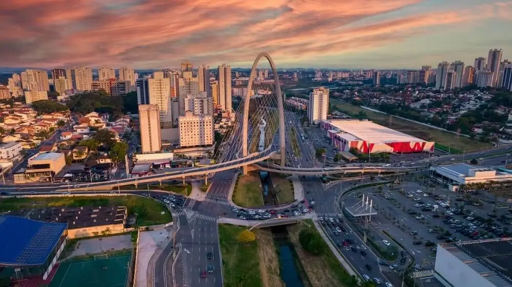 Imagem da vista aérea de São José dos Campos mostra ponte estaiada localizada na cidade com diversas avenidas que se cruzam do meio para baixo da imagem. Nos cantos superiores, há vários prédios E mais ao fundo, um céu ao entardecer com nuvens avermelhadas