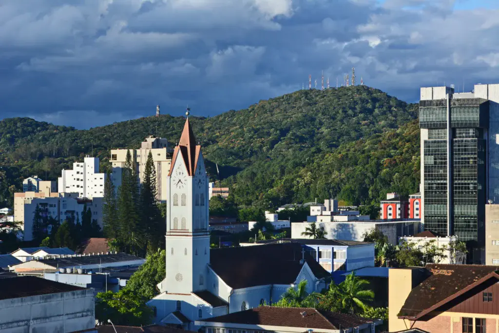 Imagem da vista aérea de Joinville mostra cenário urbano da cidade com prédios, uma igreja e vegetação ao fundo para ilustrar matéria sobre as melhores cidades de Santa Catarina