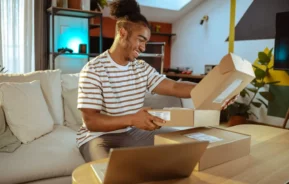 Imagem de uma pessoa sorridente sentada em um sofá abrindo uma caixa de papelão, cercada por pacotes e um laptop, em uma sala de estar moderna e aconchegante.