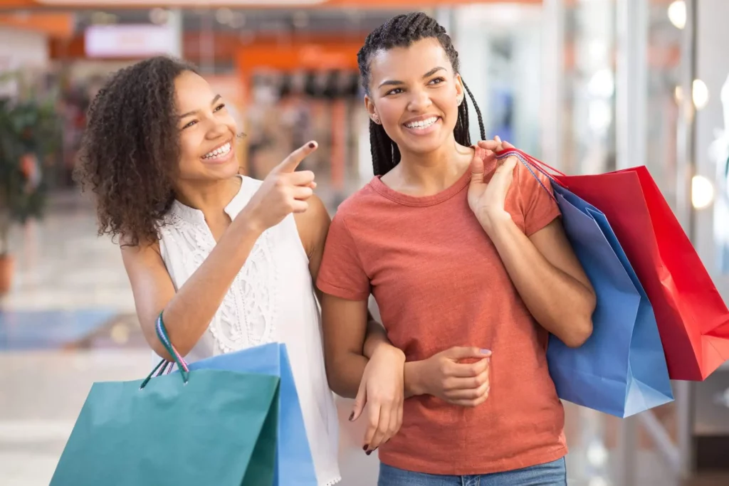 Imagem de duas mulheres sorridentes andando pelo shopping com sacolas de compras nas mãos para ilustrar matéria sobre passeio na Zona Norte de SP