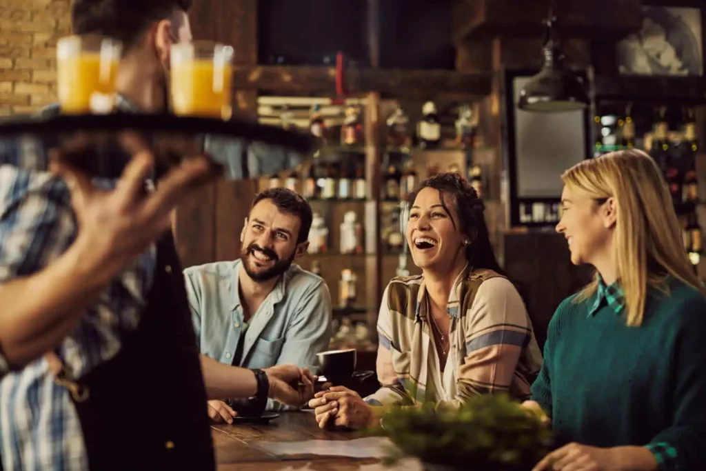 Imagem de três pessoas sentadas em um bar, sorrindo e rindo juntas enquanto um garçom carrega as bebidas. As prateleiras cheias de garrafas e copos criam uma atmosfera animada e social.