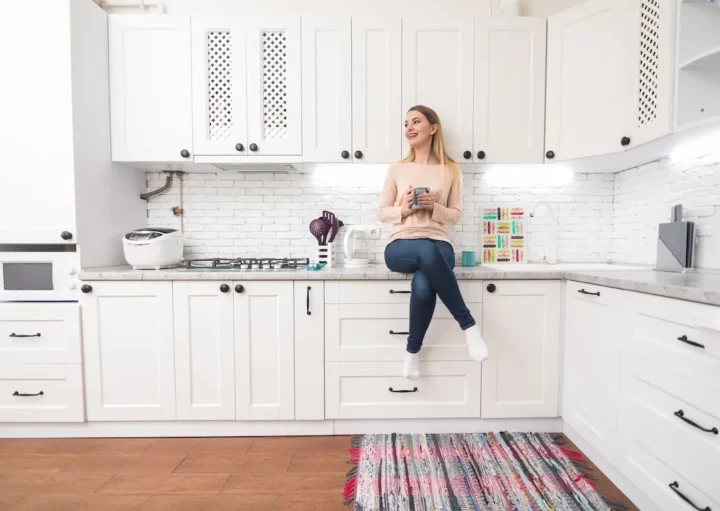 Cozinha planejada: imagem de uma mulher sentada na bancada de uma cozinha iluminada com armários brancos e piso de madeira