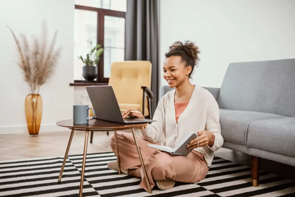 Imagem de uma jovem mulher sentada de pernas cruzadas no chão de uma sala de estar moderna, sorrindo enquanto trabalha na administração de imóveis em seu laptop em uma mesa de centro redonda. Ela segura um caderno aberto e uma caneta, com uma caneca por perto.